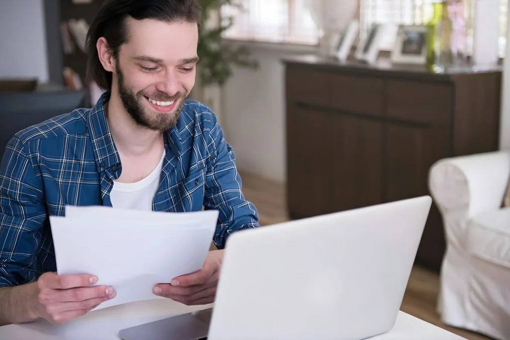 Síndico sorridente confere documentos da conta do condomínio em frente ao notebook